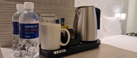 A hotel room with a wooden floor, a bed with white bedding, a black tray with a coffee maker, two water bottles, and a white mug.