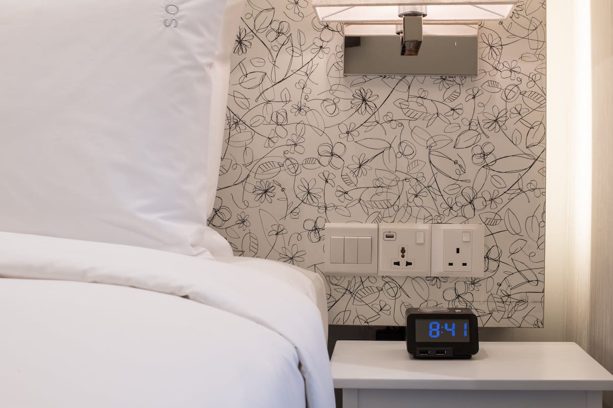 Ein ordentlich bezogenes Bett mit weißer Bettwäsche, eine digitale Uhr, die 8:41 anzeigt, und eine Wand mit einem Blumenmuster.