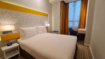 Habitación de hotel con cama, mesita de noche, lámpara y vistas al exterior a través de cortinas.