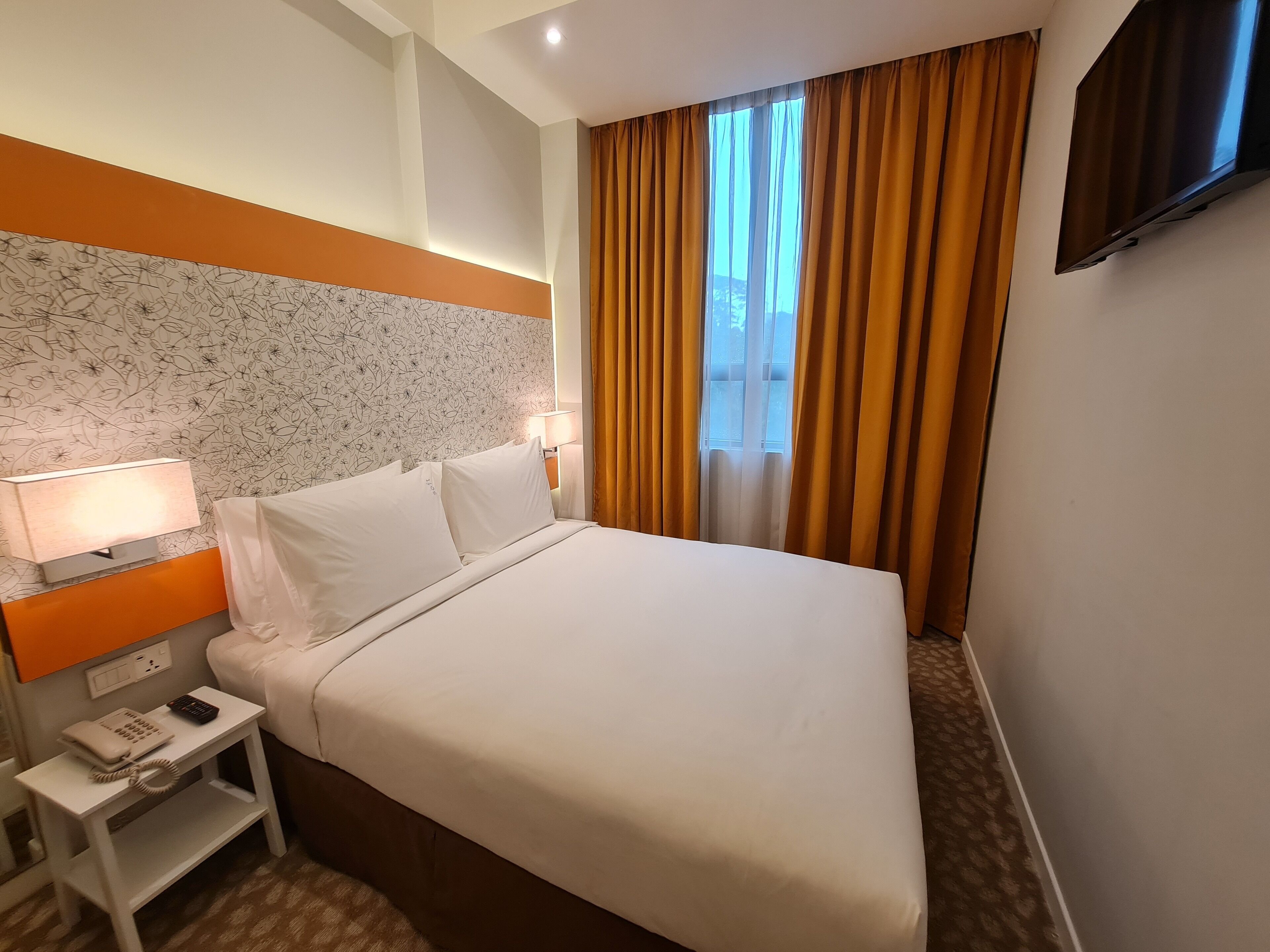 A hotel room with a bed, a nightstand with a lamp, a phone, and a TV mounted on the wall.