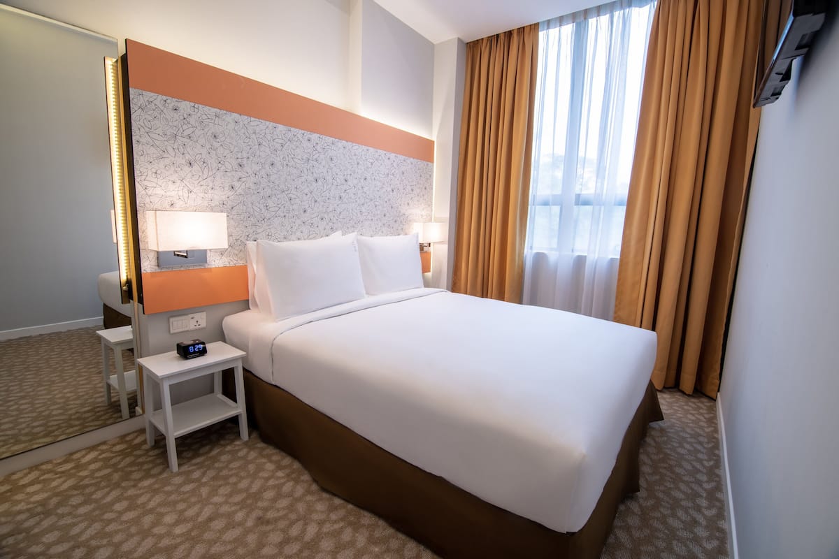 Ein Hotelzimmer mit einem großen Bett, zwei Nachttischen, einem an der Wand montierten Fernseher und einem Fenster mit Vorhängen.