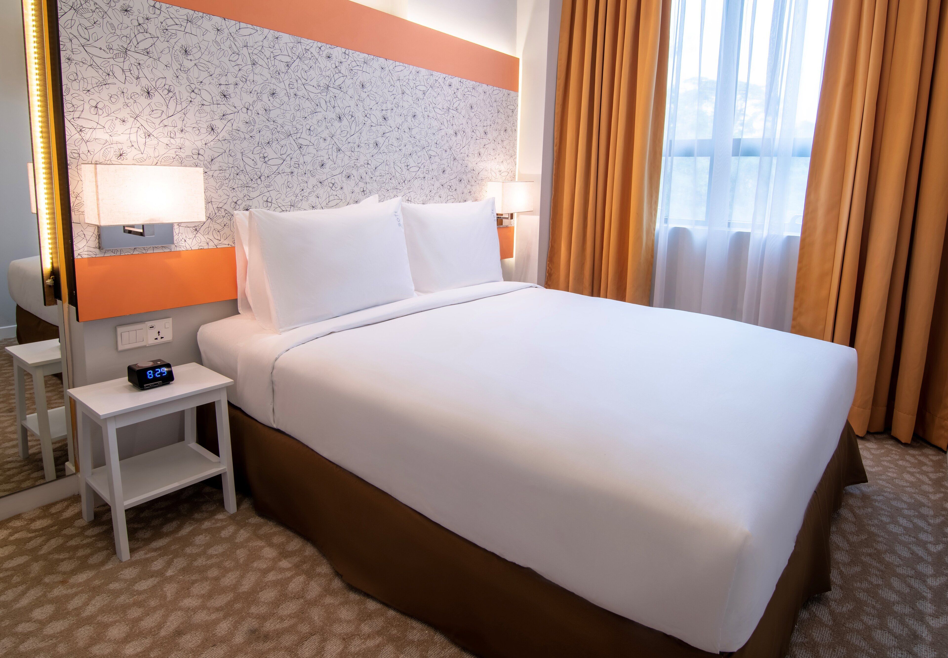 Une chambre d’hôtel avec un grand lit, une table de chevet avec une horloge, une lampe et une fenêtre avec des rideaux.