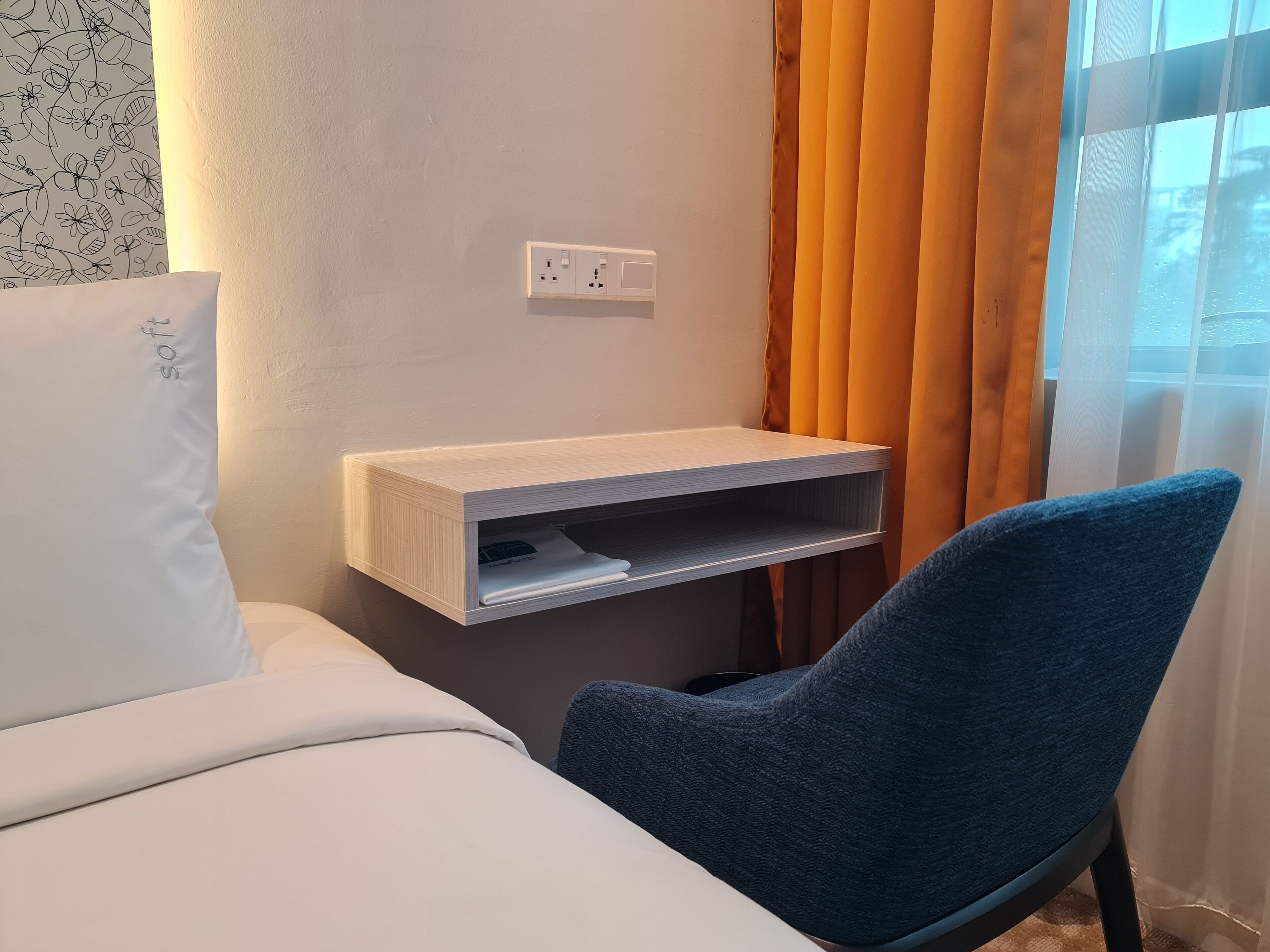 A bedroom with a bed, a blue chair, a bedside table with a lamp, and a window with curtains.