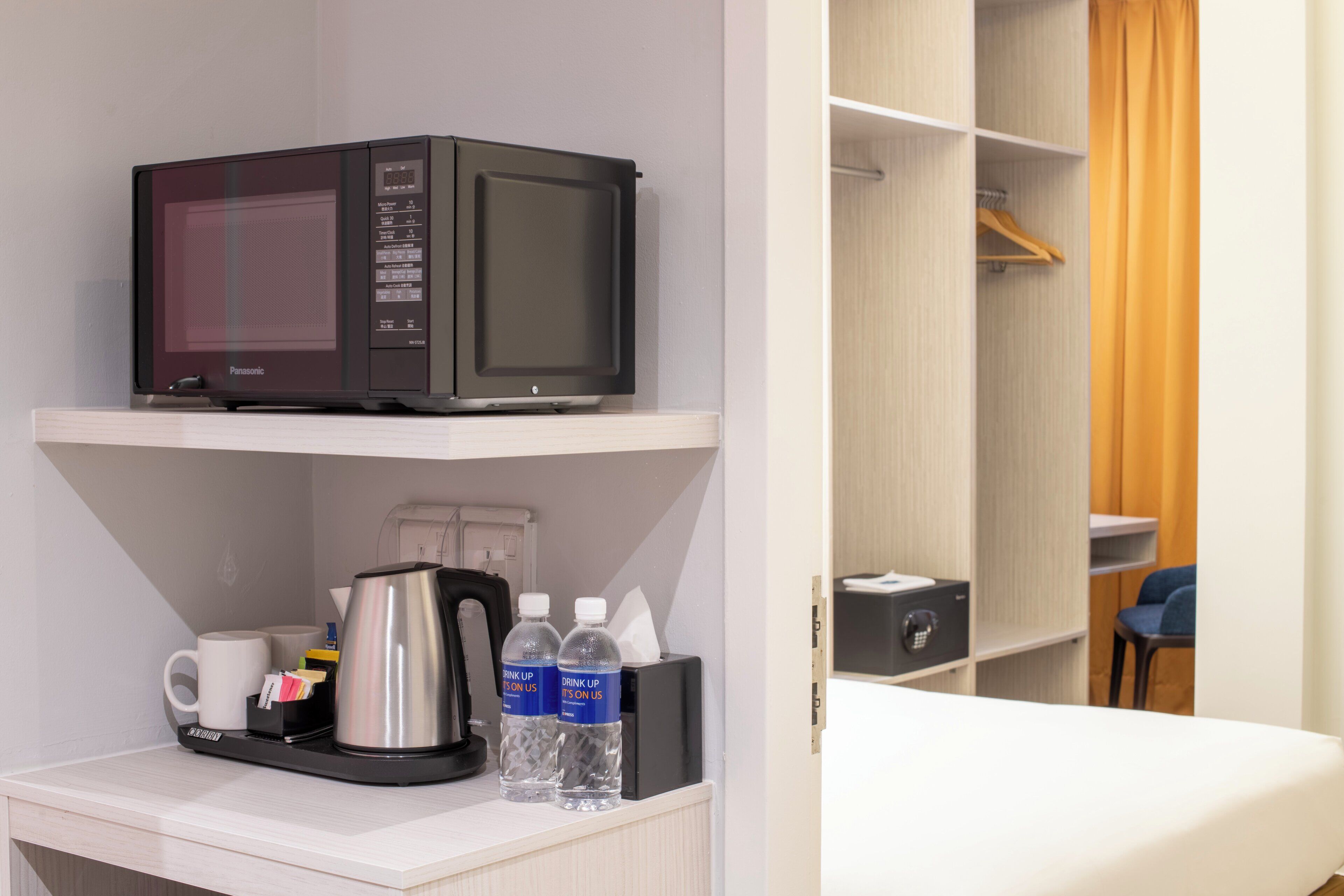 A hotel room with a microwave on a shelf, a coffee maker, two water bottles, a coffee cup, and a small table with a remote control.
