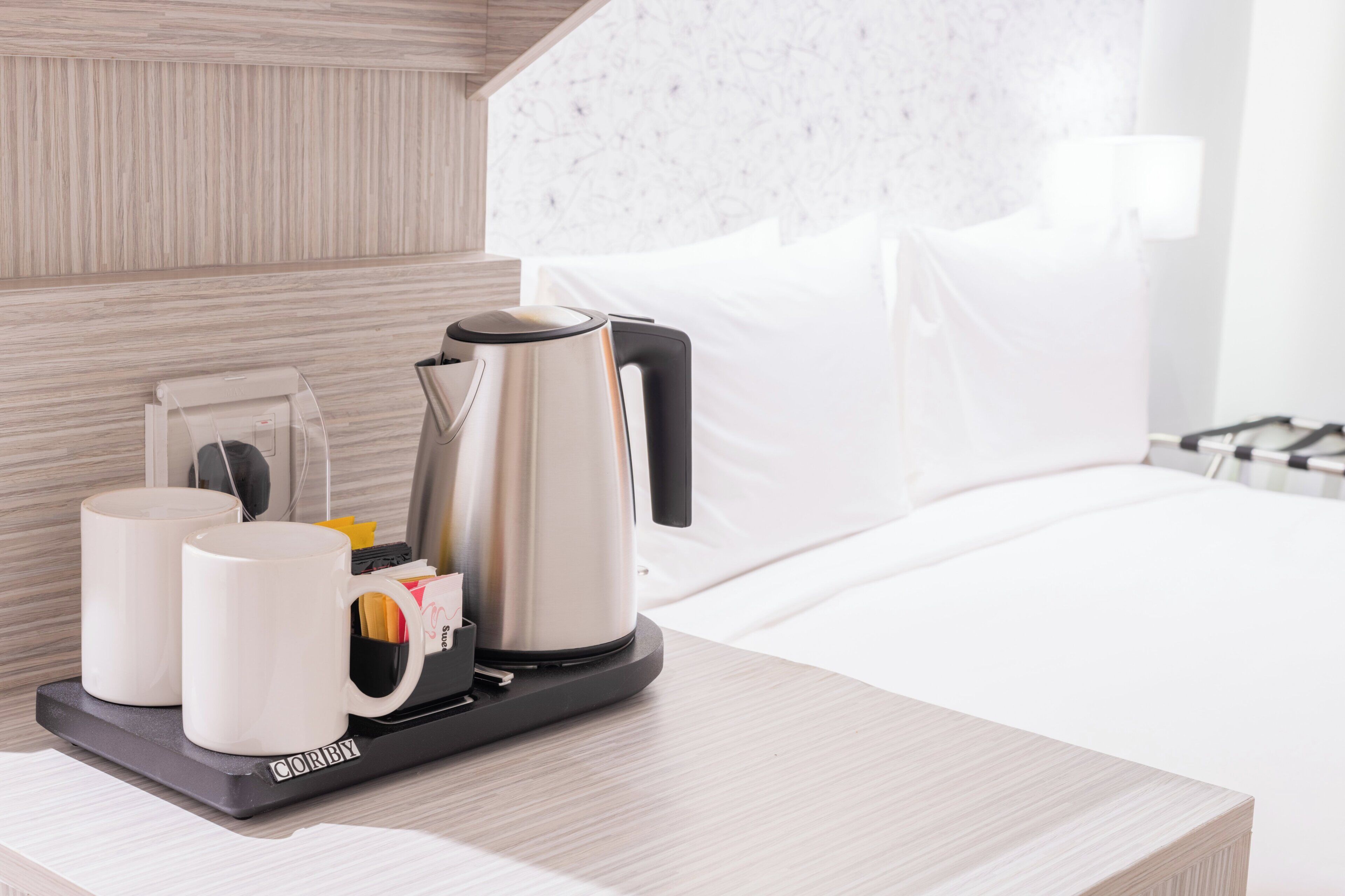 A hotel room with a bed, a bedside table with a coffee maker, two white mugs, and a tray with a bag of sugar and a spoon.