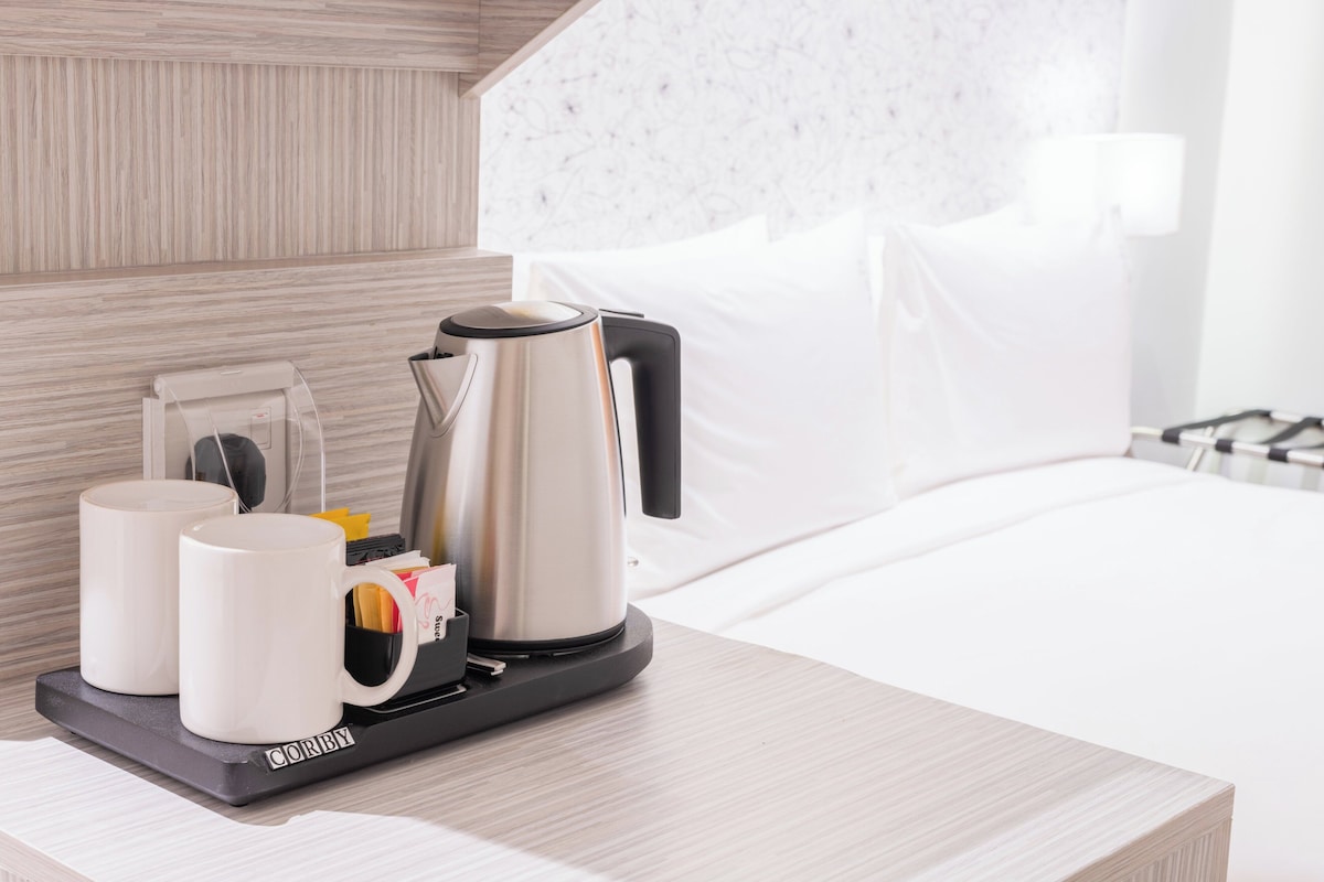 A hotel room with a bed, a bedside table with a coffee maker, two white mugs, and a tray with a bag of coffee pods.