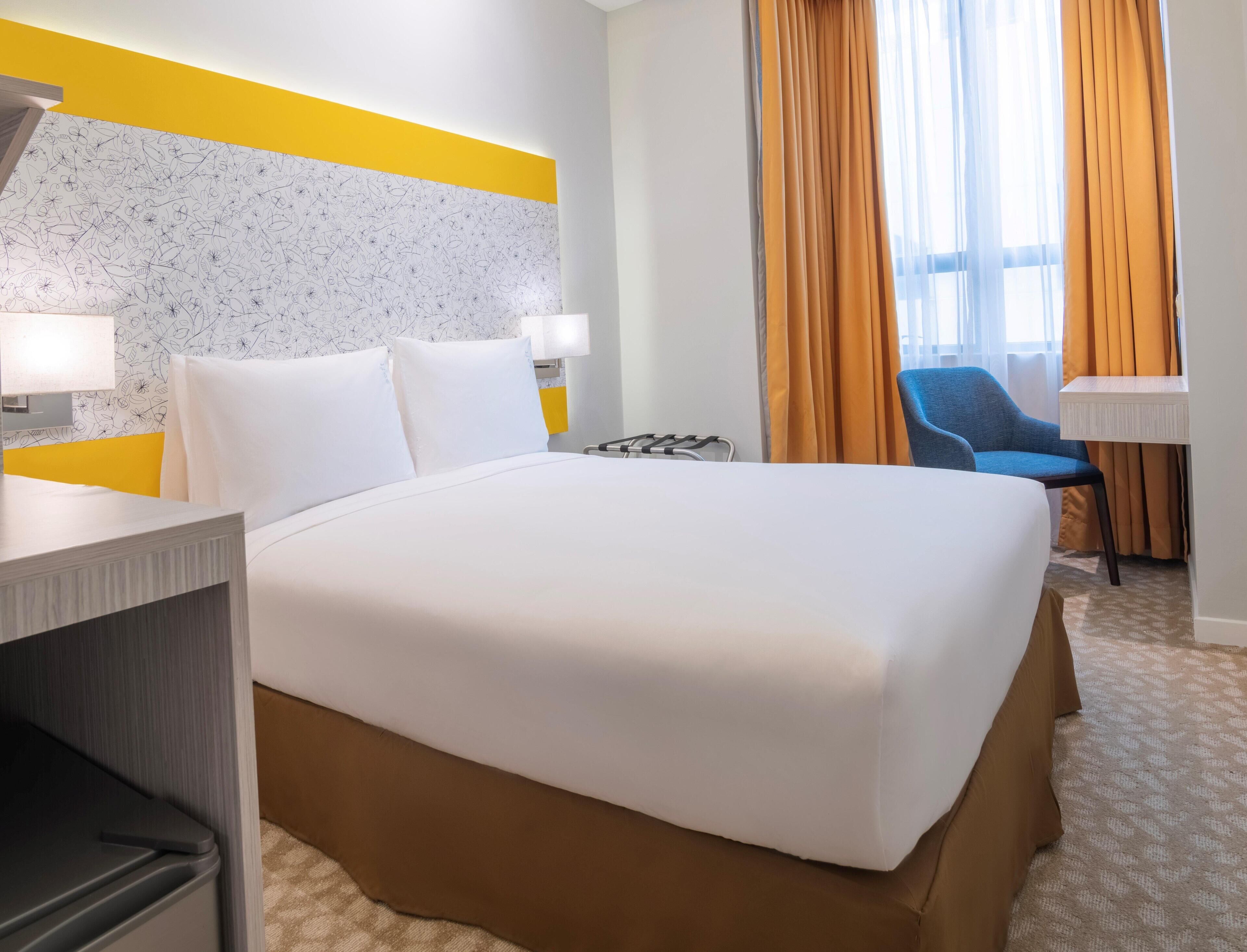 A hotel room with a large bed, two bedside tables, a desk, a chair, and a window with curtains.