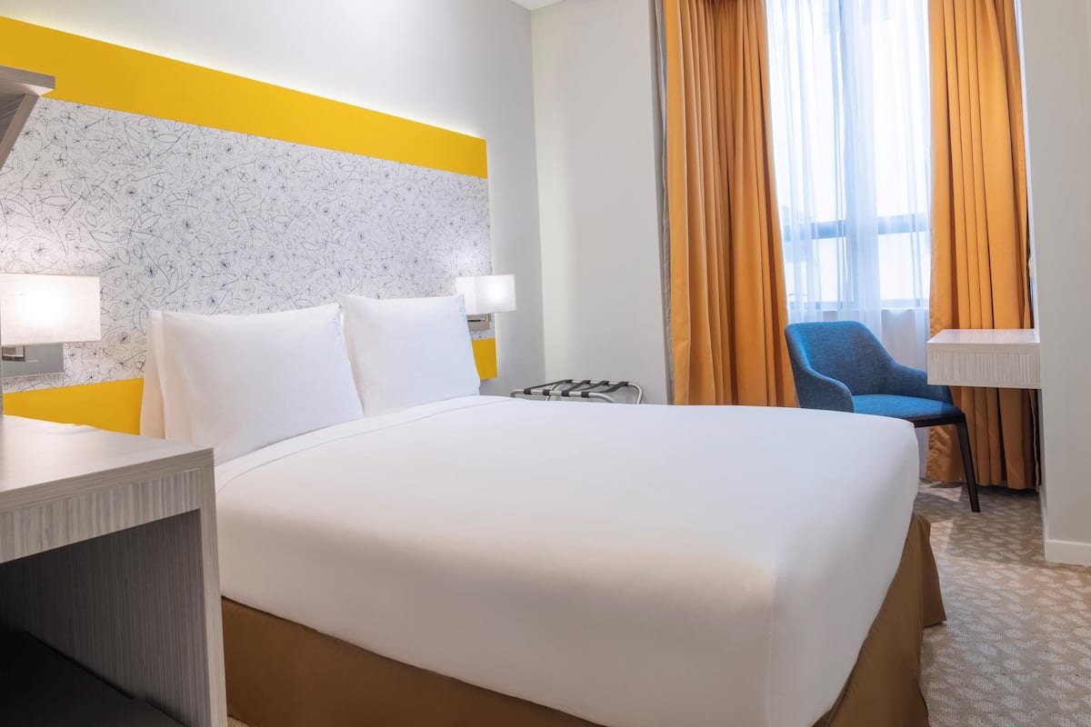 Quarto de hotel com uma cama grande, duas mesas de cabeceira, uma escrivaninha, uma cadeira e uma janela com cortinas.