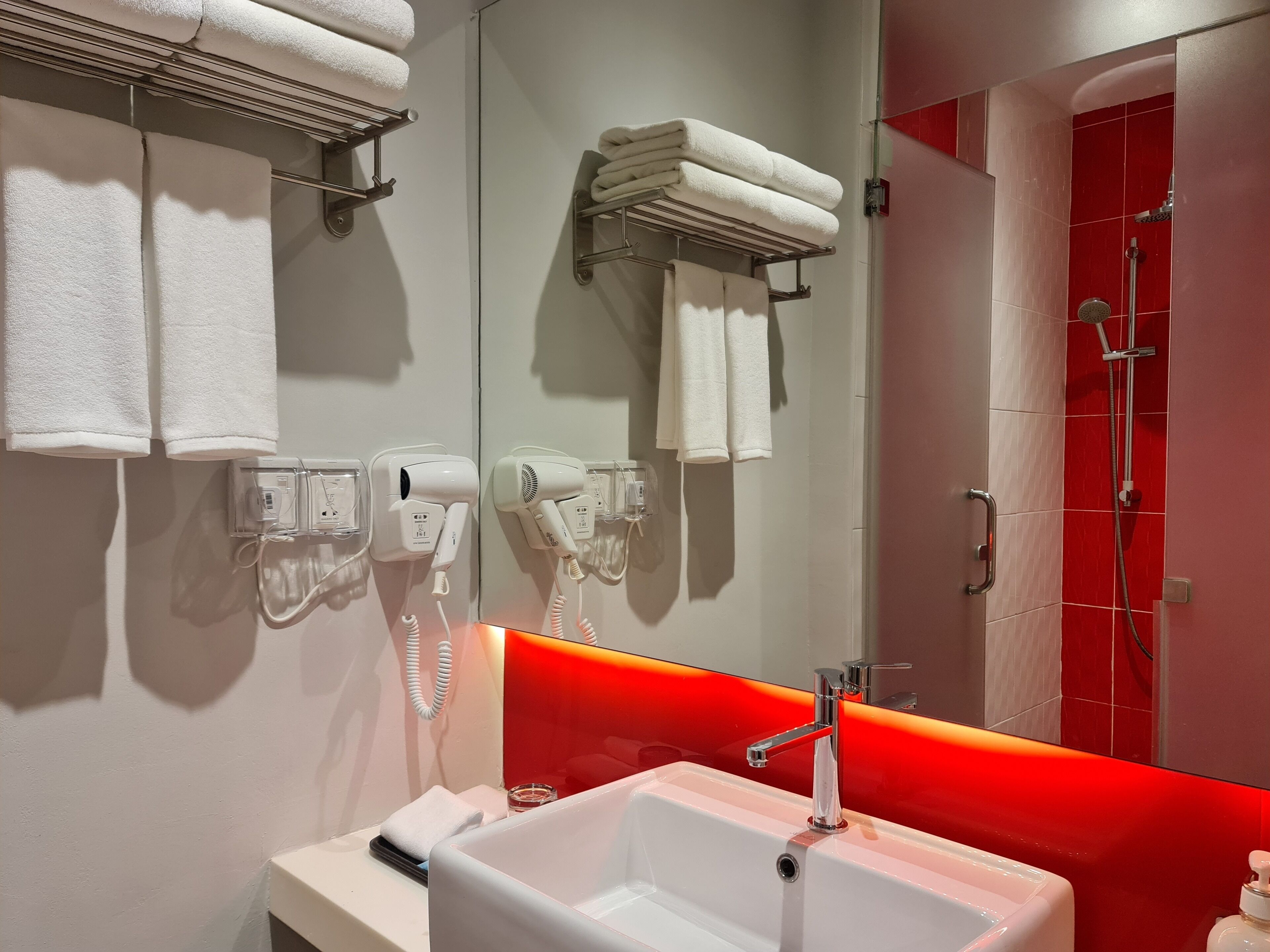A modern bathroom with a white sink, two hair dryers mounted on the wall, and a red-tiled shower area.