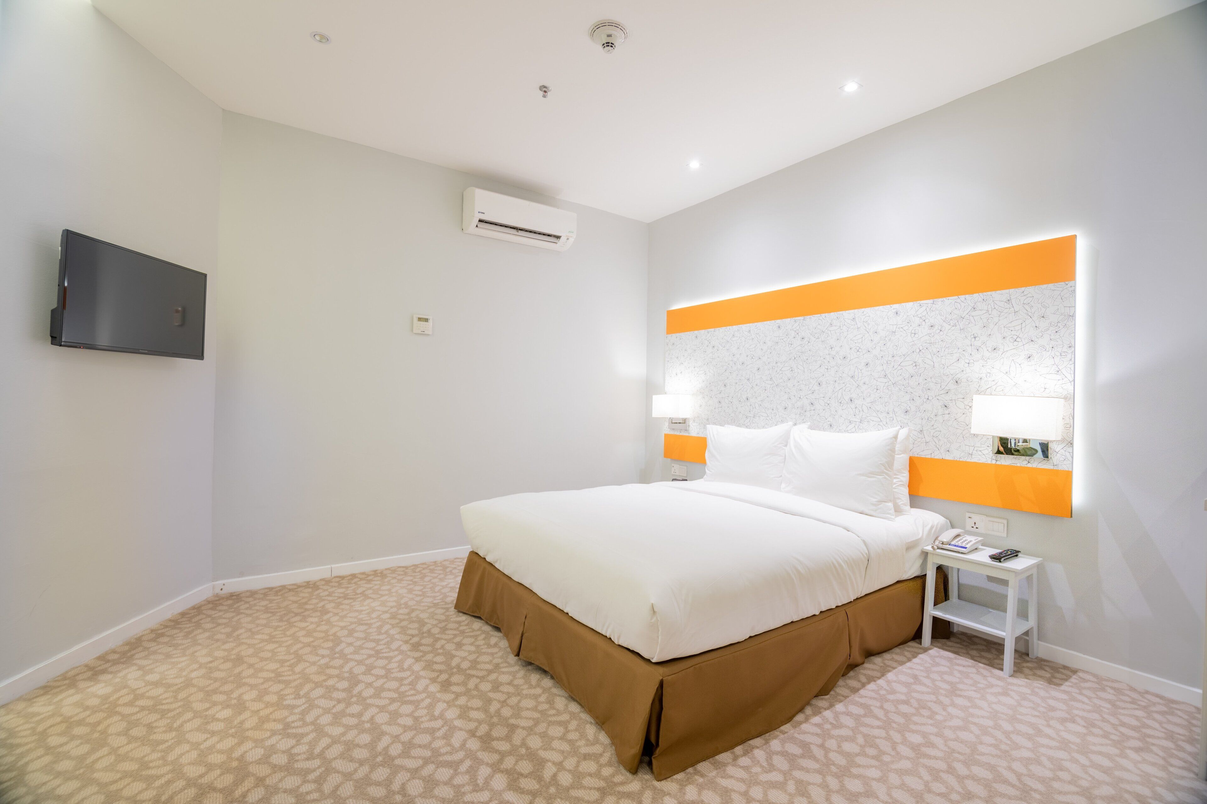 A hotel room with a bed, a TV mounted on the wall, an air conditioning unit, a bedside table with a lamp, and a patterned carpet.