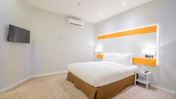A hotel room with a bed, a TV mounted on the wall, an air conditioning unit, a bedside table with a lamp, and a patterned carpet.