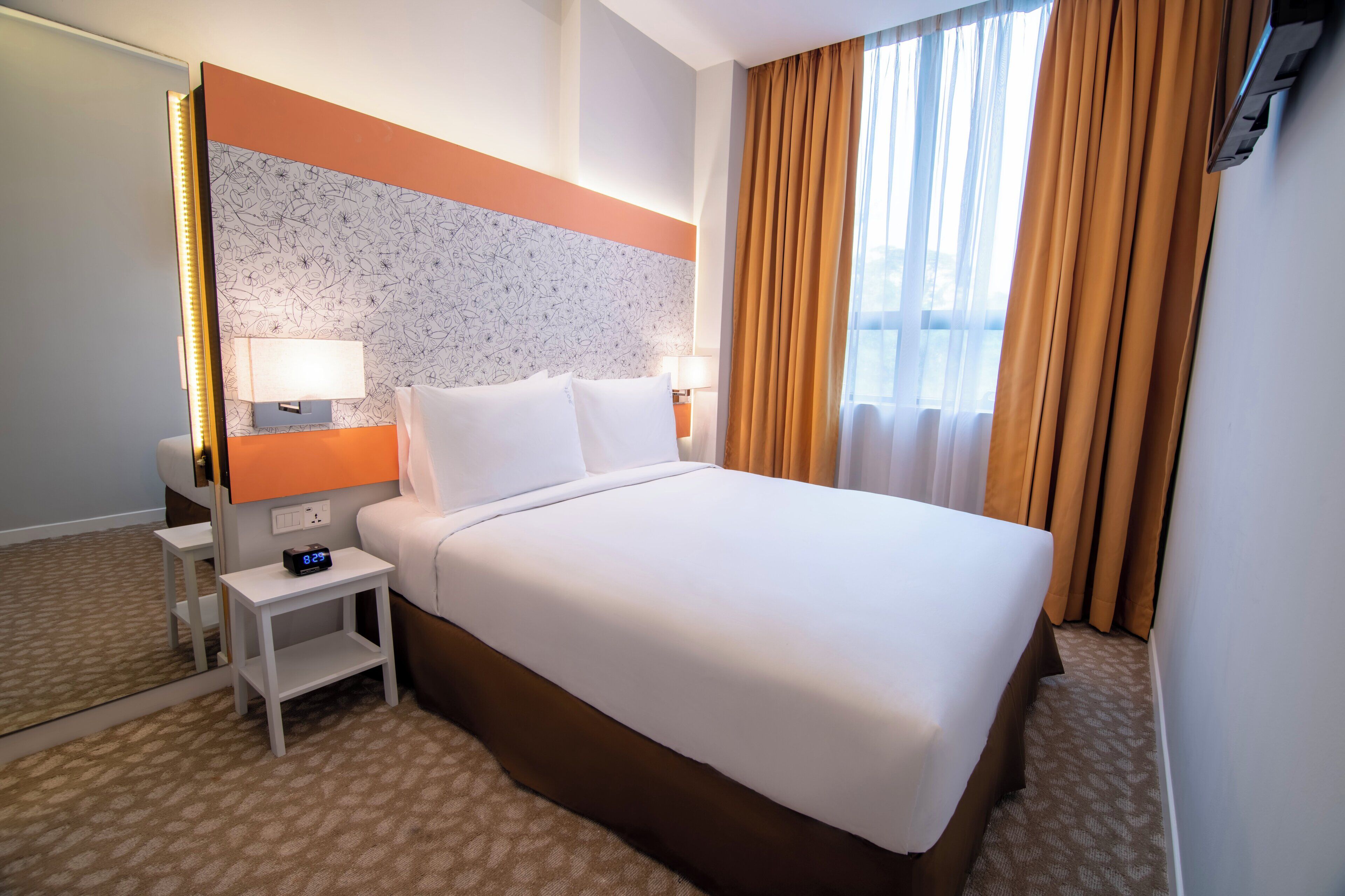 Una camera d'albergo con un letto grande, un comodino con sveglia digitale, una TV a muro e una finestra con tende.
