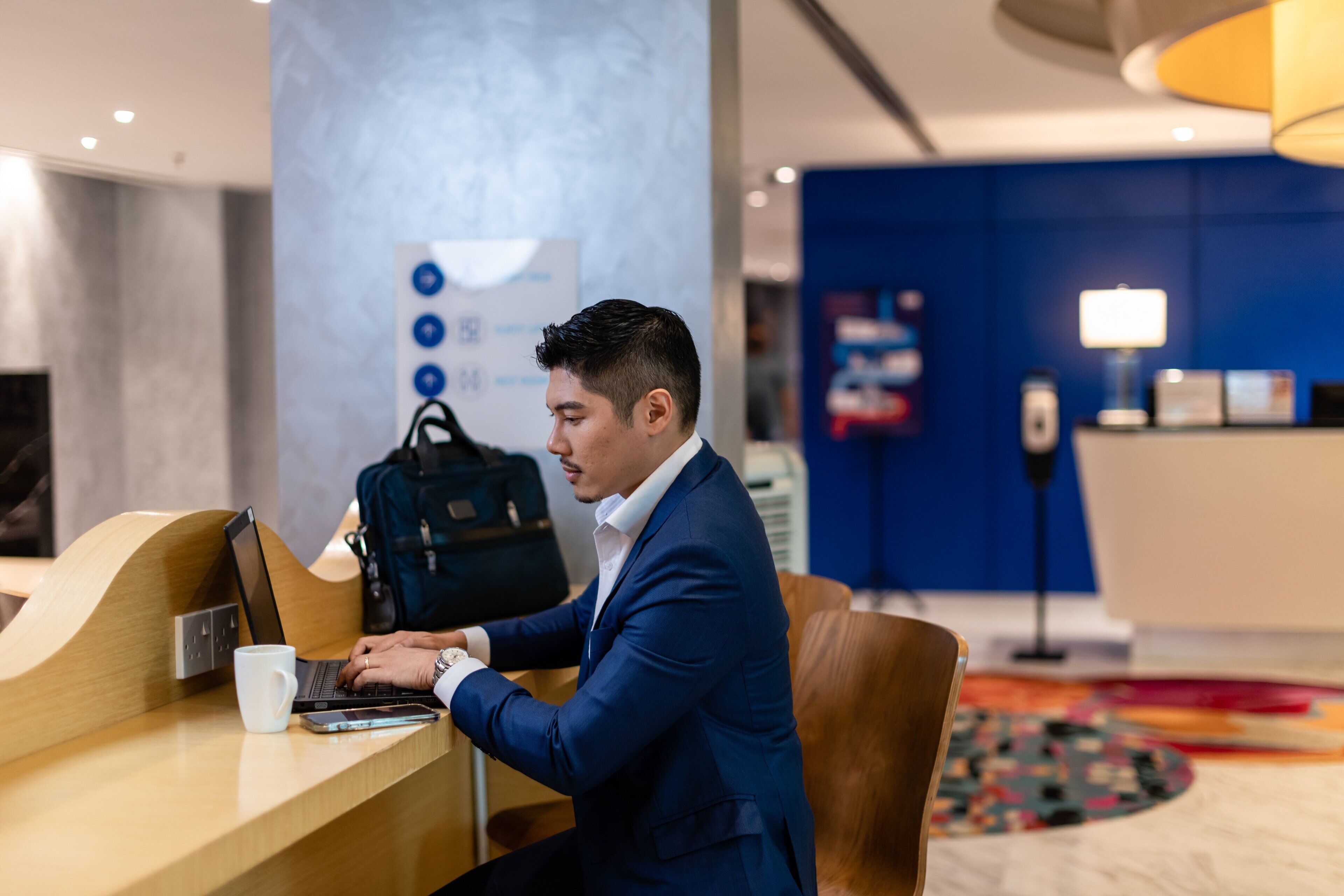A man in a blue suit working on a laptop at a reception desk in a modern hotel lobby.