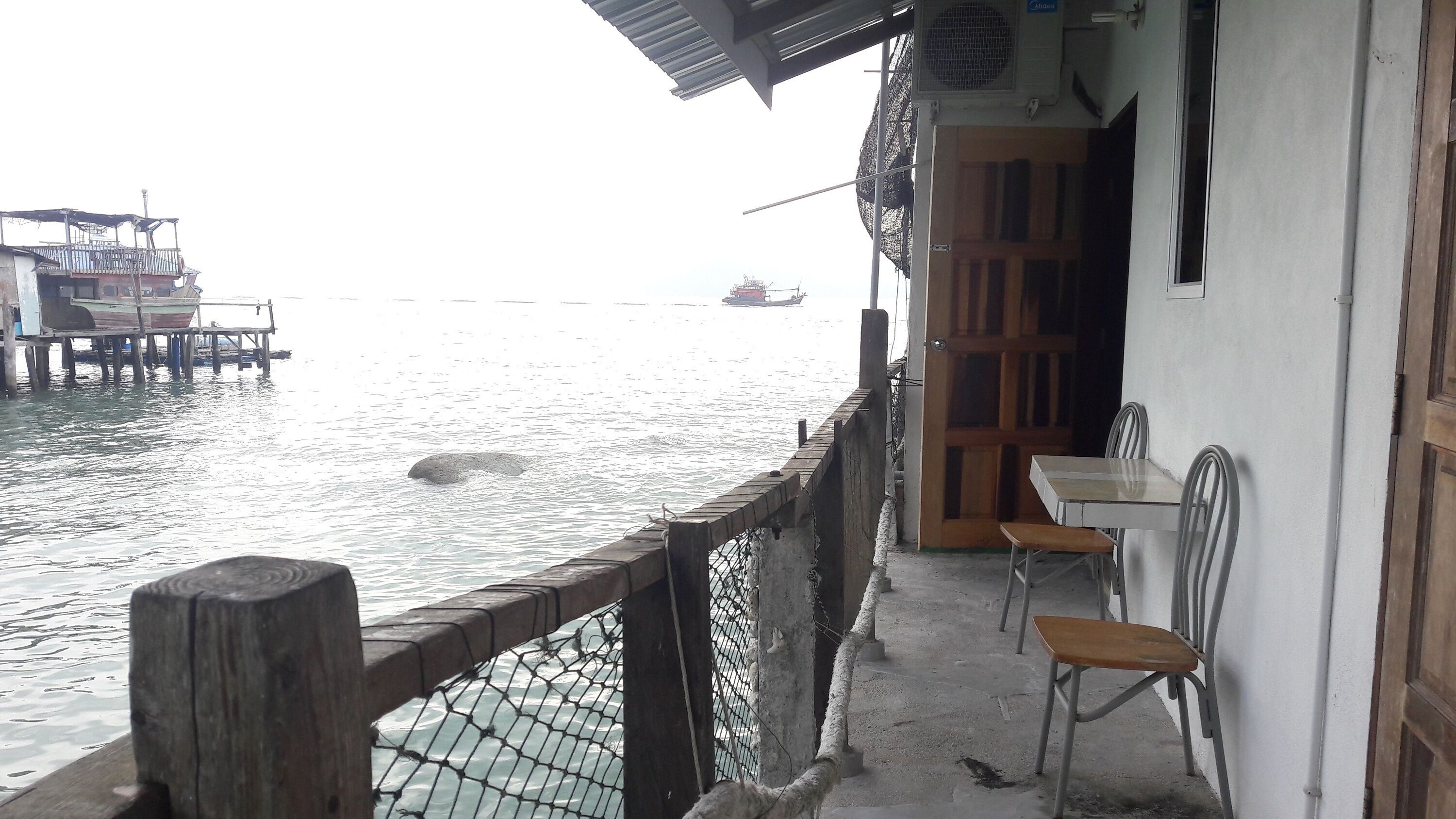 A wooden pier with a netted railing, a table and chairs on the right, and a boat in the water.