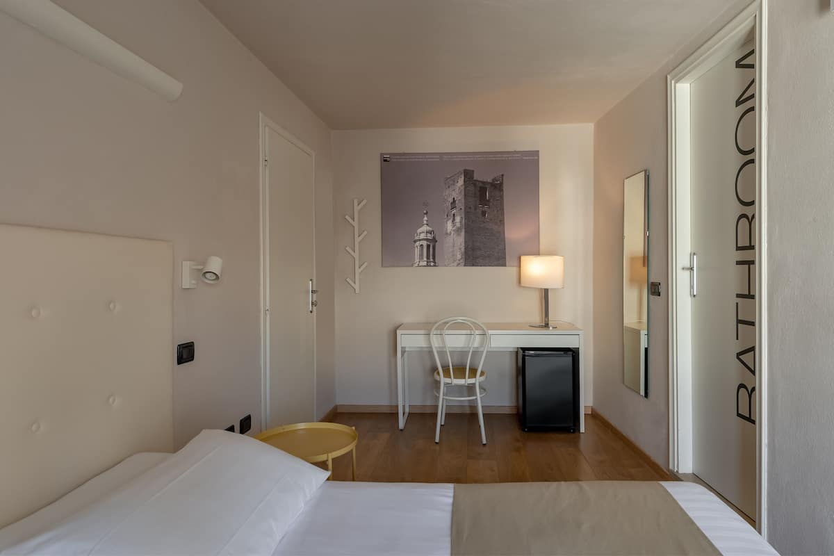A hotel room with a bed, a desk with a chair, a small table, a lamp, and a picture on the wall.