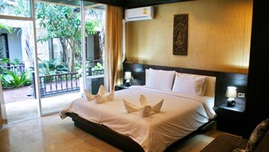 A hotel room with a large bed, two bedside tables with lamps, a wall-mounted air conditioner, and a view of a garden through a sliding glass door. - Victorian Samui Condominium (Koh Samui)