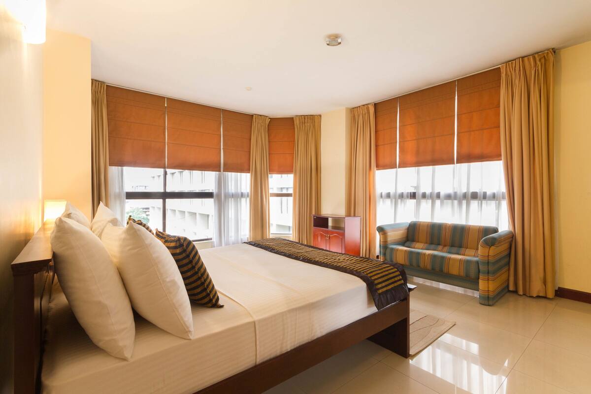A hotel room with a bed, a sofa, a desk, and large windows with curtains.
