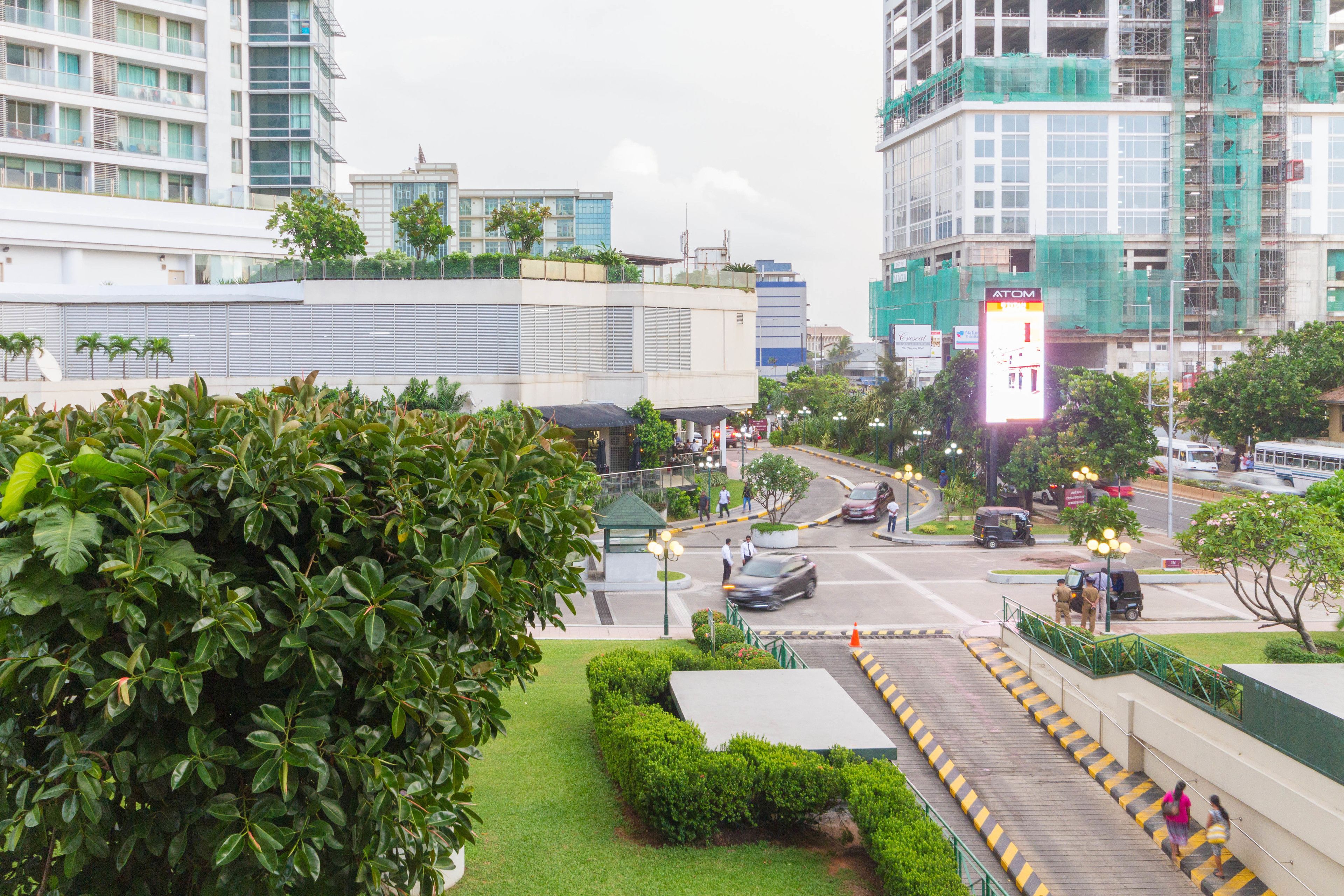 Un paysage urbain moderne, caractérisé par un mélange de bâtiments résidentiels et commerciaux, un espace vert bien entretenu et une intersection animée où circulent véhicules et piétons.