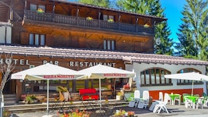 A two-story wooden building with a restaurant on the ground floor. There are outdoor tables and chairs under umbrellas. - Hotel Vecchia America (Dimaro Folgarida)
