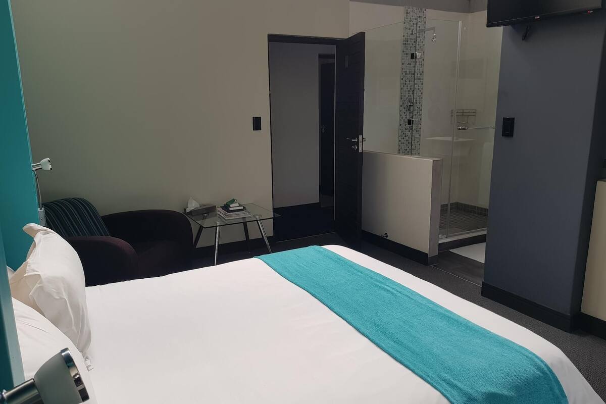 Ett hotellrum med en säng, ett skrivbord, en stol, ett badrum med dusch och en tv som är monterad på väggen.
