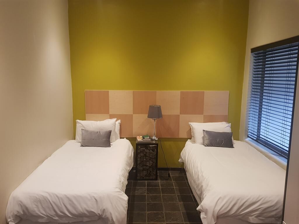 A hotel room with two beds, a headboard with a checkered pattern, a nightstand with a lamp, and a window with blinds.