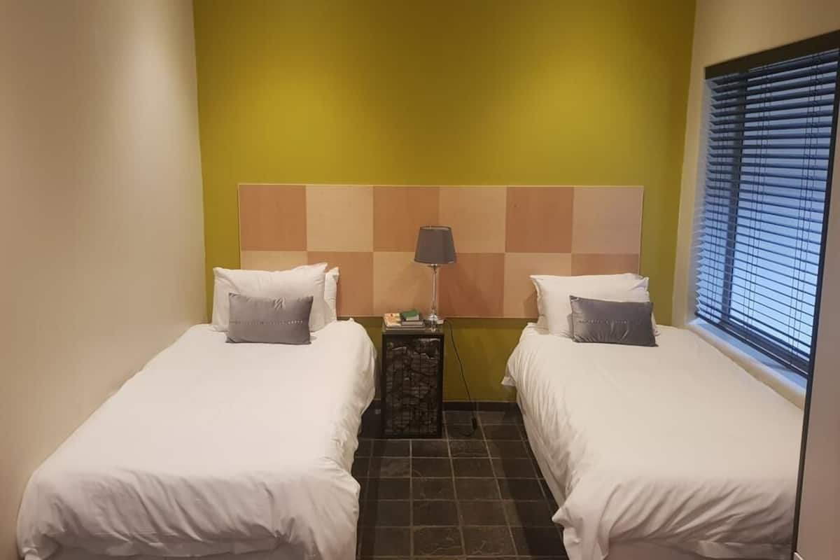 Habitación de hotel con dos camas, cabecera de estampado de cuadros, mesita de noche con lámpara y ventana con persianas.