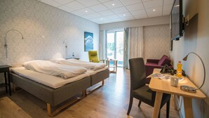 A hotel room with a bed, a desk with a chair, a table with a bottle and a cup, a lamp, and a TV on the wall. - Almaas Hotell Stord  (Stord)