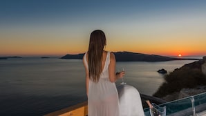 A woman in a white dress standing by a railing, overlooking a sunset over water with distant landforms. - Elite Luxury Suites (Santorini)