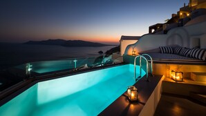A modern infinity pool with a glass railing, a cozy seating area with striped cushions, and a view of the sea at sunset. - Elite Luxury Suites (Santorini)