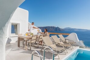 A sunlit outdoor dining area with a pool and lounge chairs, overlooking a clear blue sea and distant mountains. - Elite Luxury Suites (Santorini)