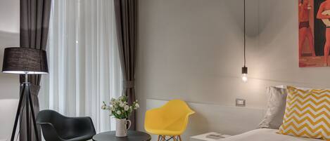 Habitación de hotel moderna con una cama, una mecedora negra, una silla amarilla, una pequeña mesa con un florero y un cuadro en la pared.