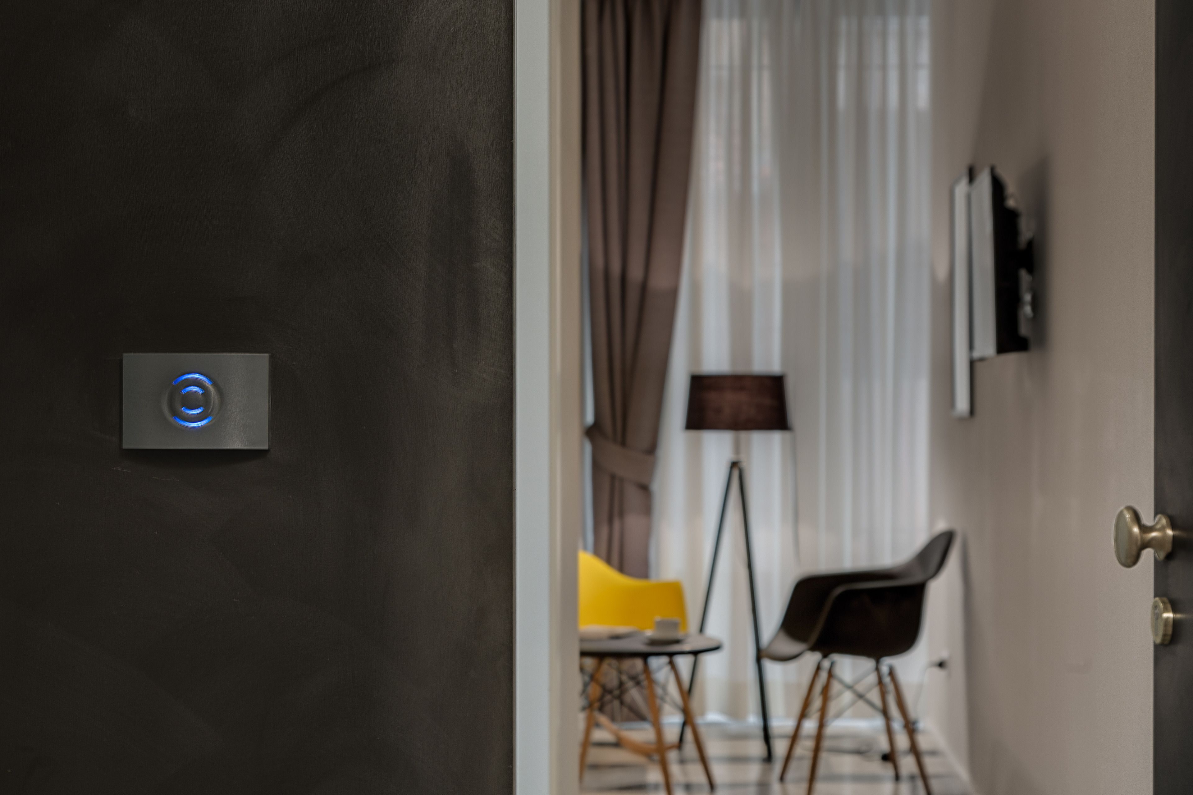 A modern interior with a door featuring a blue illuminated sensor, a dark wall, a lamp, and a chair.