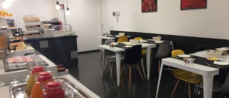 A modern dining area with tables and chairs, a buffet counter with food items, and two portraits on the wall.