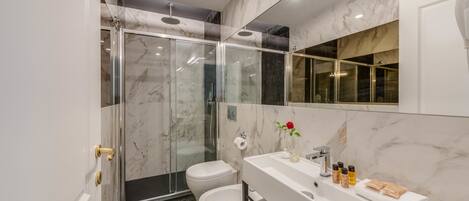 Un baño moderno con ducha envuelta en vidrio, un lavamanos blanco con artículos de aseo y un salpicadero de mármol.