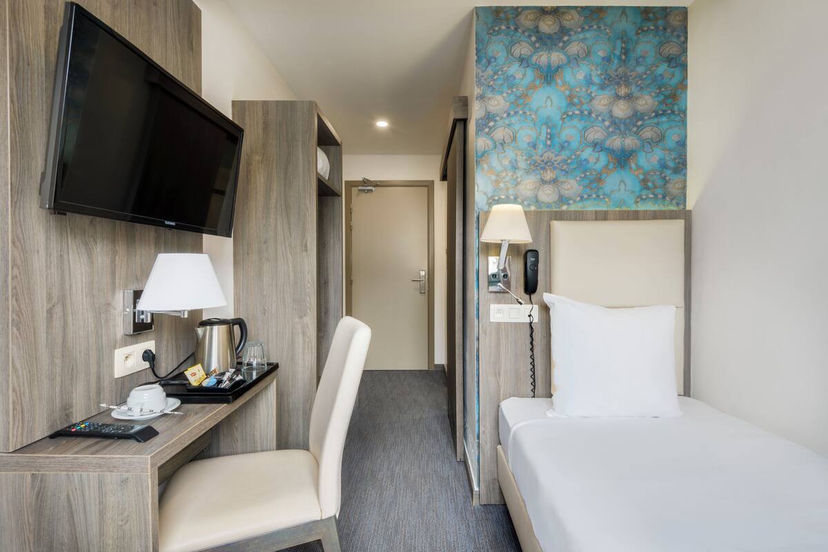 Habitación de hotel con televisor de pantalla plana, escritorio con lámpara, cafetera, silla, cama con almohada y pared con diseño.