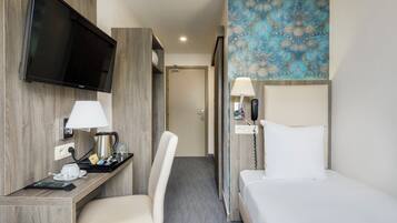 Quarto de hotel com televisão de tela plana, mesa com luminária, cafeteira, cadeira, cama com travesseiro e parede estampada.