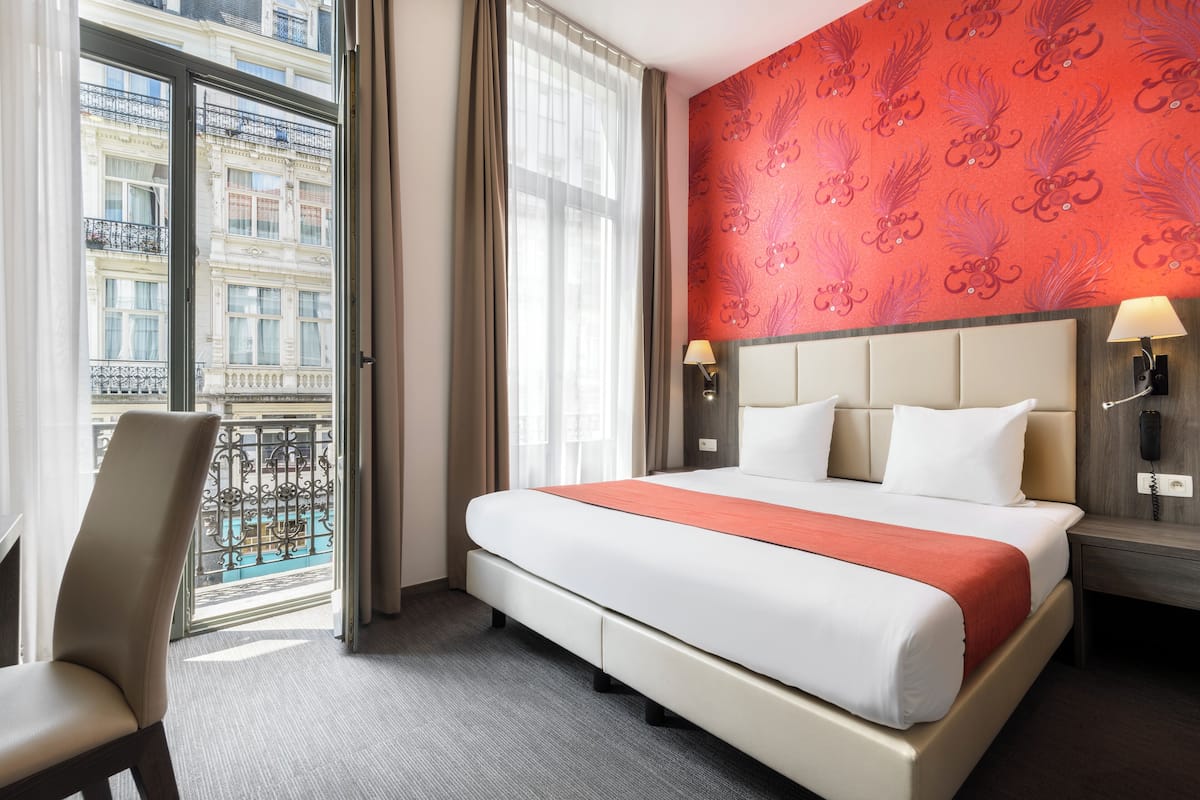 Habitación de hotel con una cama grande, una silla, una mesita de noche con lámpara y vista a un edificio a través de una ventana.