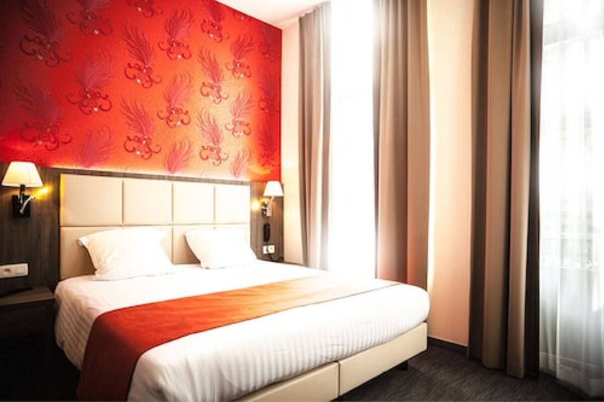 Habitación de hotel con cama, dos lámparas de noche, pared con diseño rojo y una ventana con cortinas.
