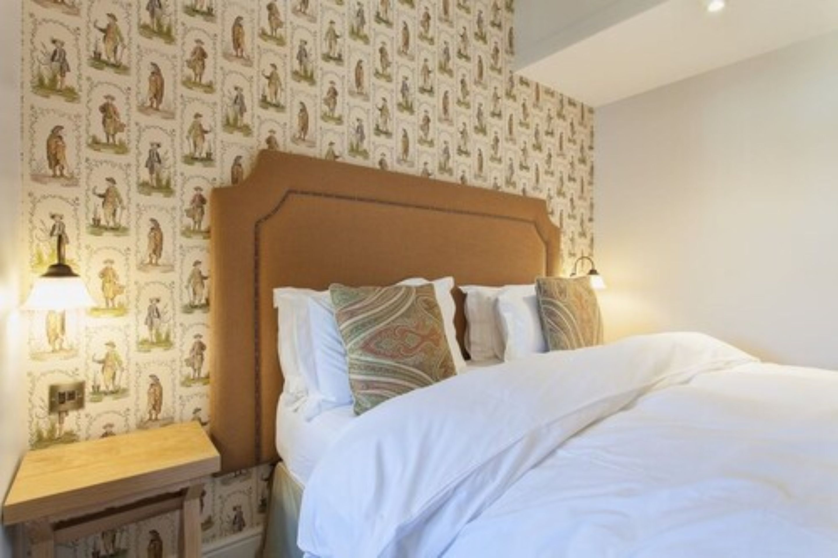Una cama bien tendida con sábanas blancas y almohadones estampados, una mesita de noche de madera con una lámpara, y una pared cubierta con un papel tapiz que repite figuras históricas.