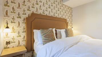 Una cama bien tendida con sábanas blancas y almohadones estampados, una mesita de noche de madera con una lámpara, y una pared cubierta con un papel tapiz que repite figuras históricas.