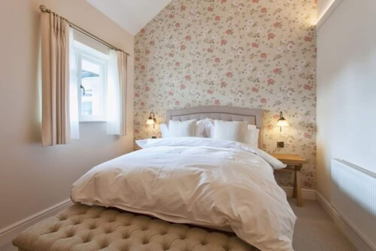 Quarto com cama, janela com cortinas, mesa de cabeceira com abajur e papel de parede floral.