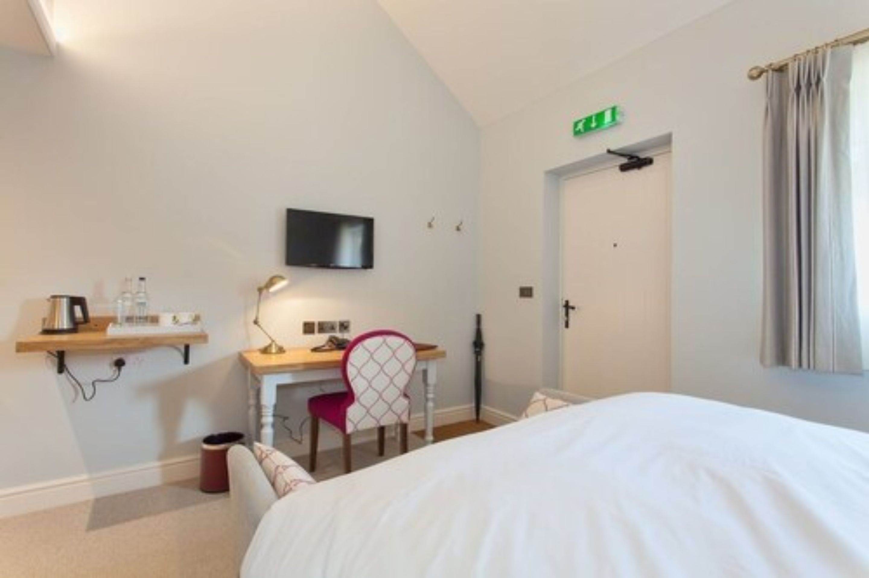 A hotel room with a bed, a desk with a chair, a television, and a door with an emergency exit sign.