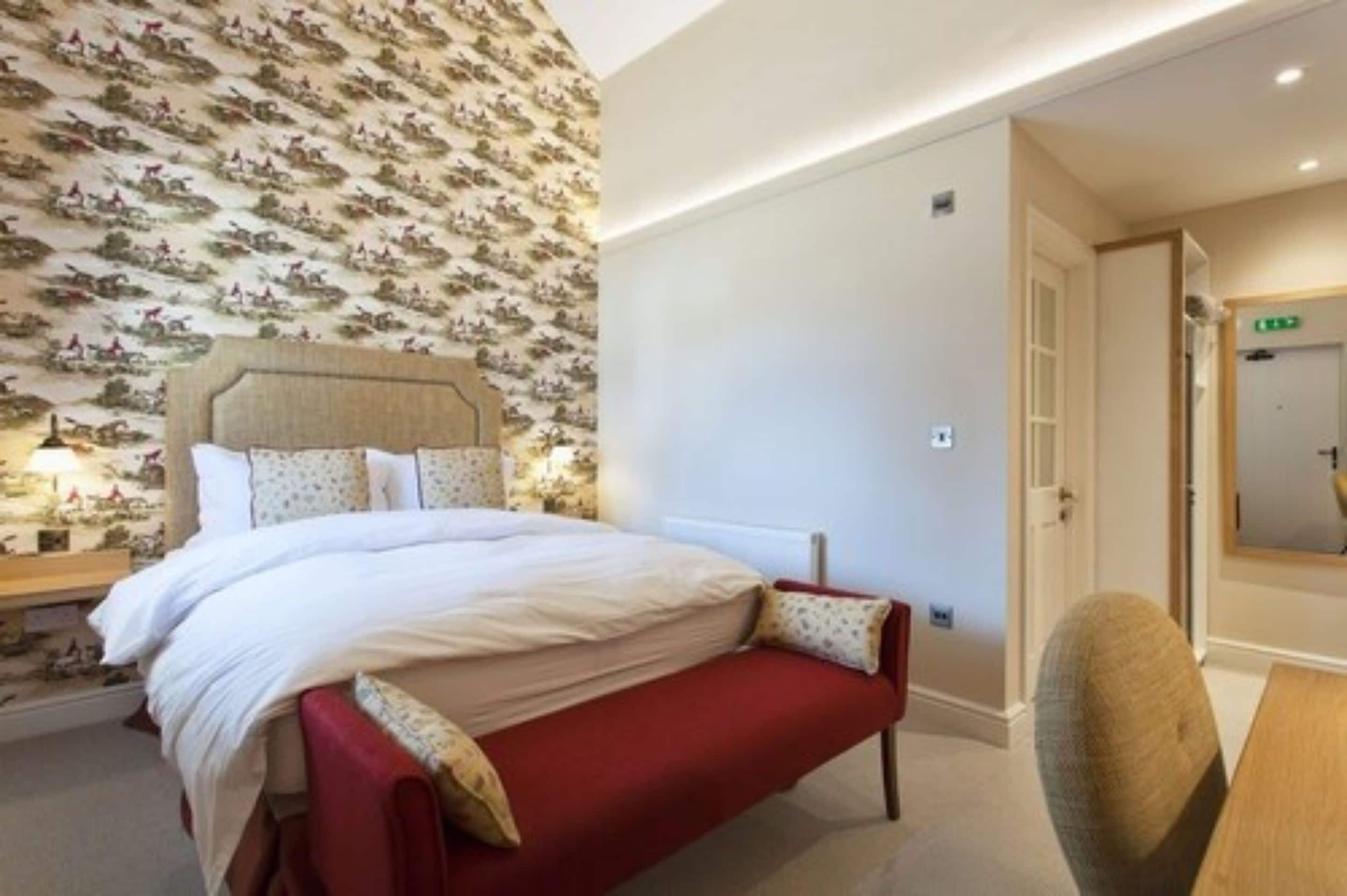 Une chambre d’hôtel bien aménagée, avec un grand lit, une table de chevet avec une lampe, un banc rouge et un mur recouvert d’un papier peint à motifs.