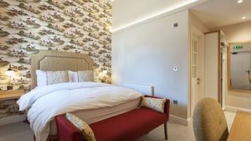 Une chambre d’hôtel bien aménagée, avec un grand lit, une table de chevet avec une lampe, un banc rouge et un mur recouvert d’un papier peint à motifs.