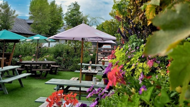 An outdoor patio area with picnic tables and umbrellas, surrounded by lush greenery and flowering plants. - The Inn West End (Woking)