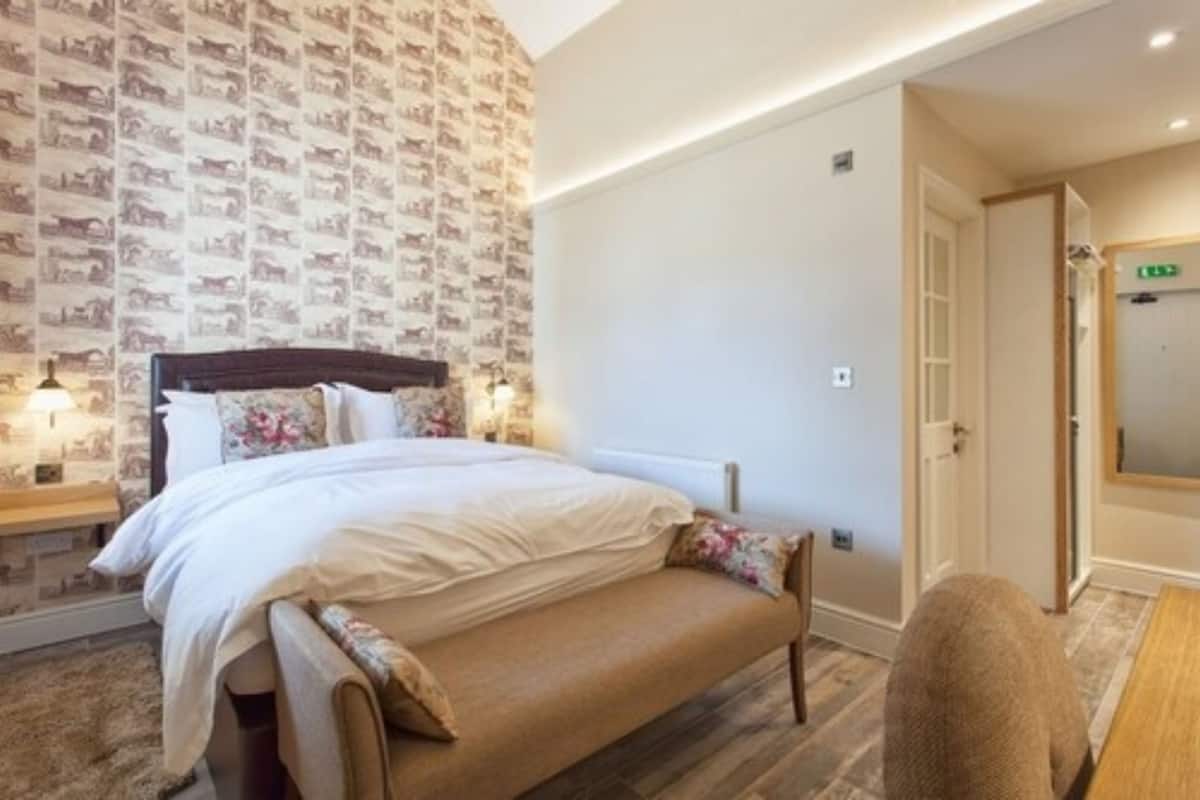 Quarto com cama, um banco, mesa de cabeceira e parede com papel de parede estampado.