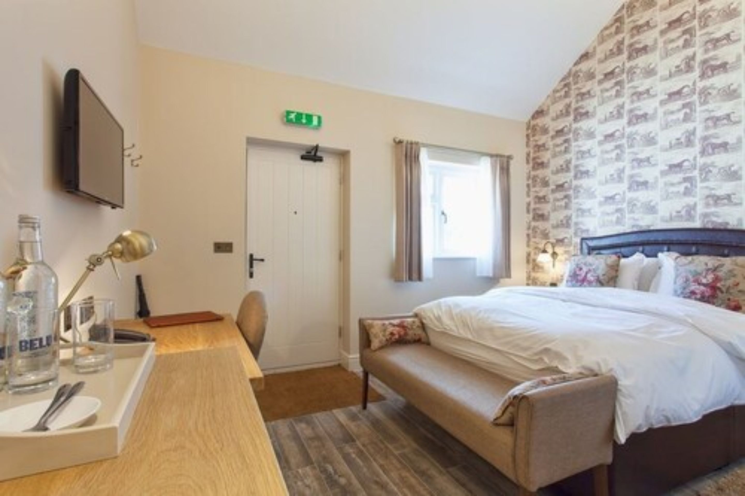 A hotel room with a bed, a desk with a lamp, a chair, a bedside table with glasses and a bottle, and a patterned wallpaper.