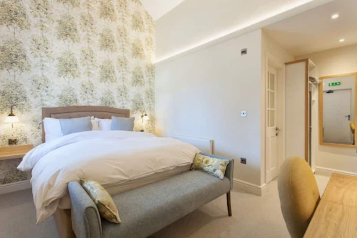 Quarto com cama, mesas de cabeceira, um banco, espelho e porta.