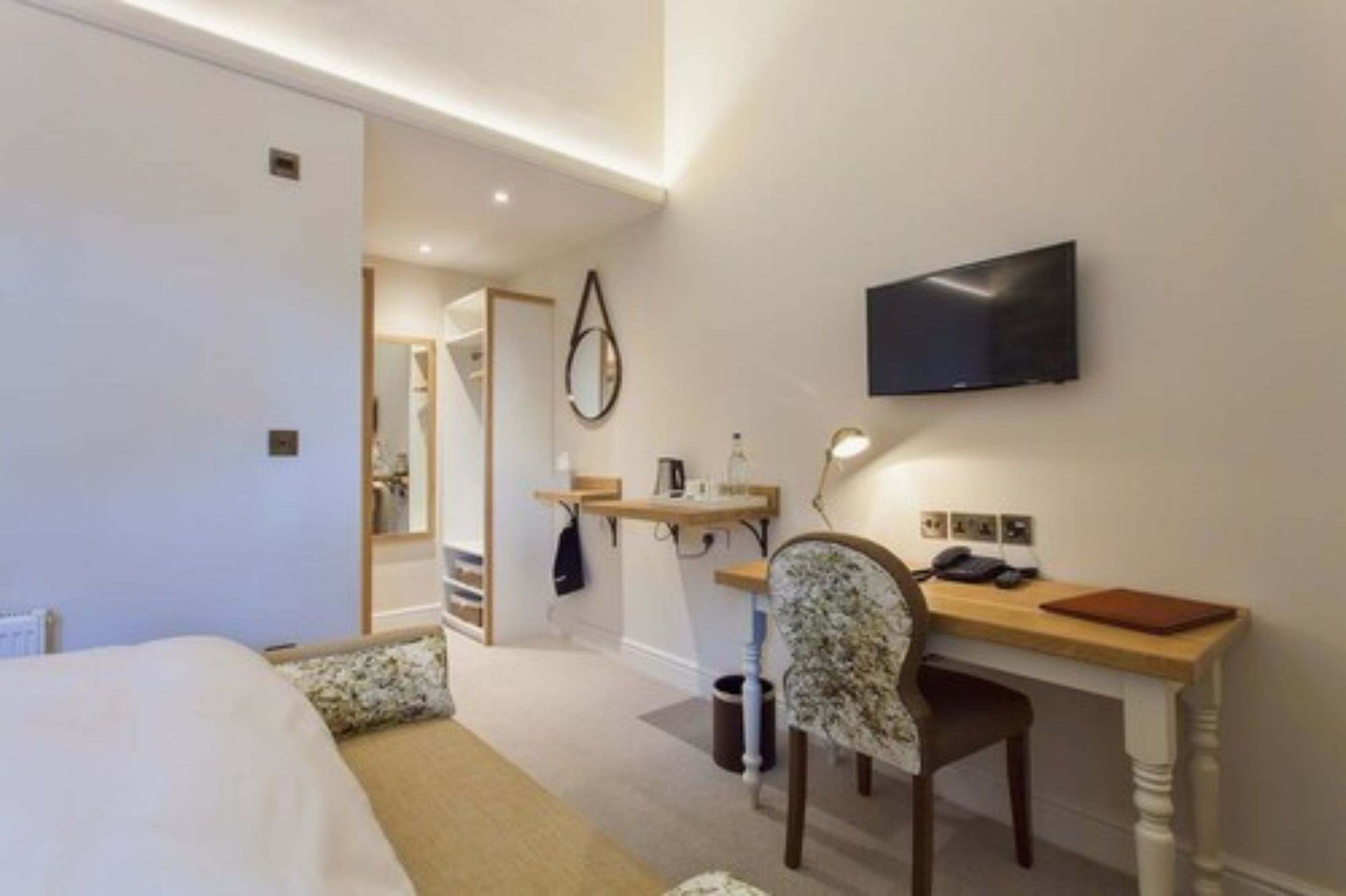 Ein Hotelzimmer mit einem Bett, einem Schreibtisch mit Stuhl, einem an der Wand montierten Fernseher und einem kleinen Regal mit Gegenständen.