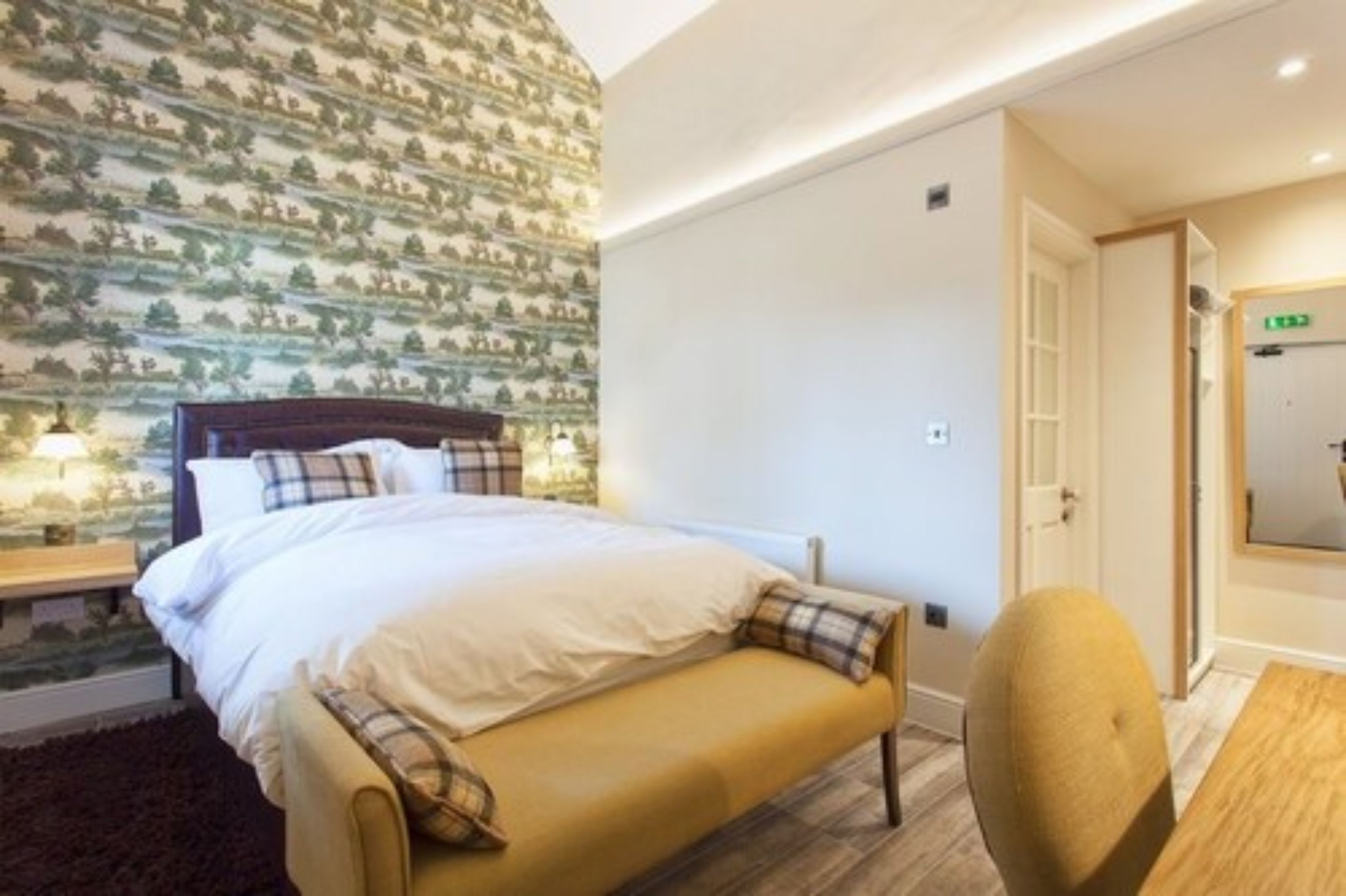 A bedroom with a bed, a bench, a chair, a nightstand, and a wall with a tree pattern.